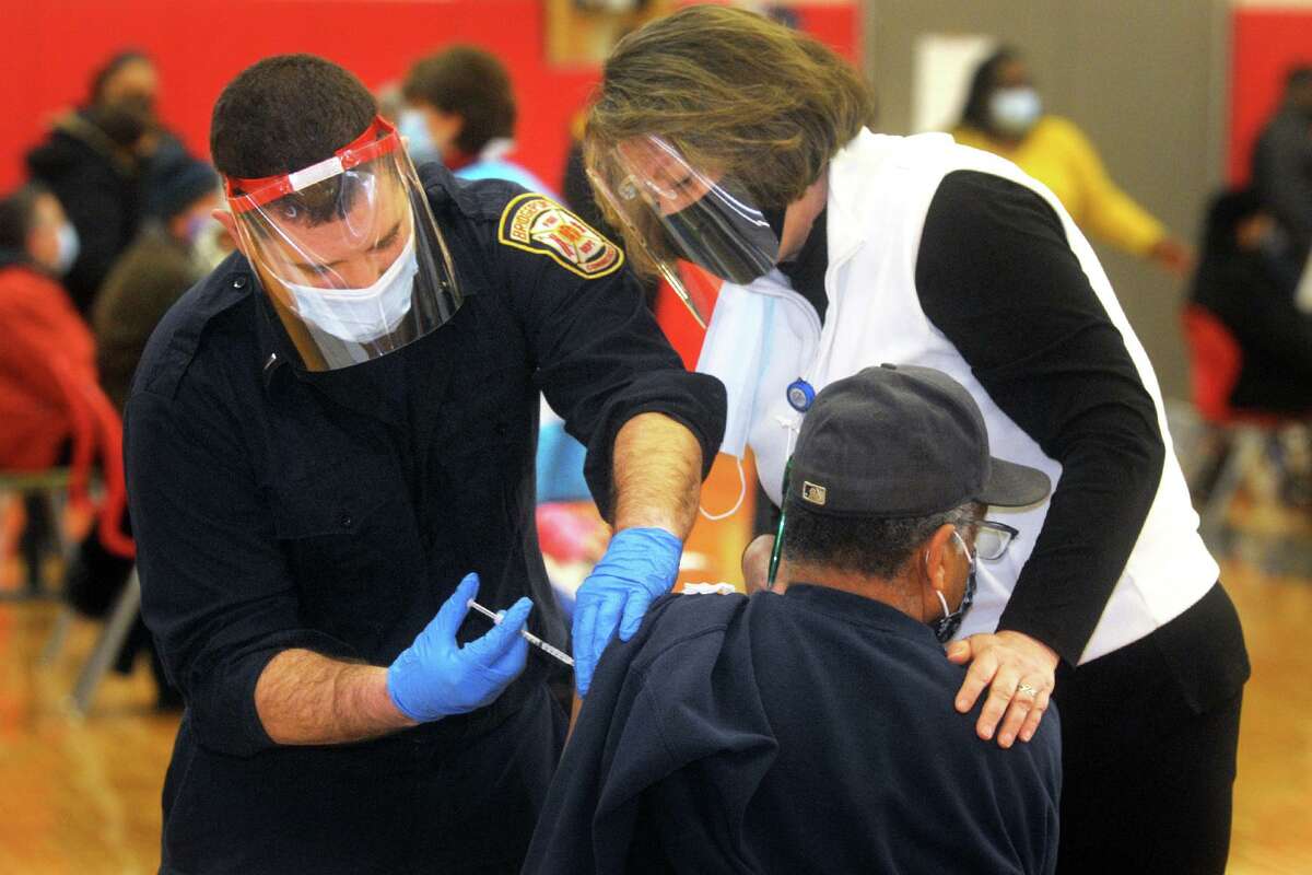 Nurse Elin Loh assists Lt. Ken Benedict of the Bridgeport Fire Department as he administers a COVID-19 vaccination to a man at the weekly vaccination clinic held in the gymnasium of Central High School, in Bridgeport, Conn. Feb. 10, 2021.