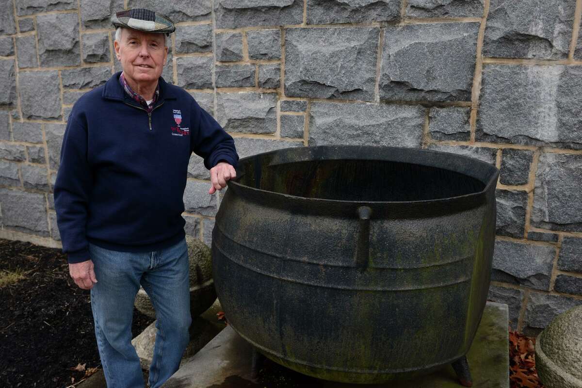 Jack Walsh, former head of the Derby Historical Society, poses next to an iron kettle on display next to the Derby Public Library, in Derby, Conn. Dec. 29, 2021. The kettle was used for boiling down whale blubber in the 1800’s. Walsh has created the Historic Birmingham Borough District Map, an online interactive map including many points of historic interest around Derby, Connecticut’s smallest city, including the Derby Public Library, built in 1912.