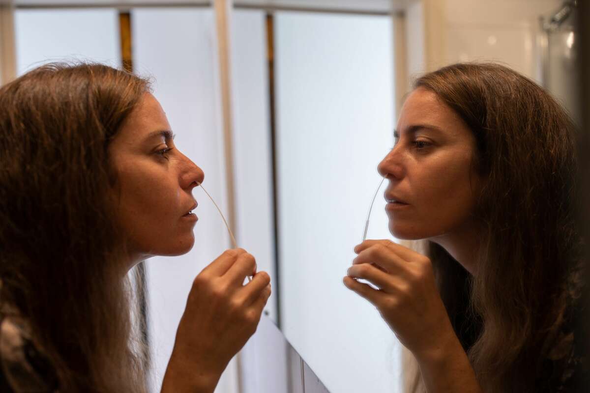 A woman performs a COVID-19 self-test via nasal swab in front of bathroom mirror at home.