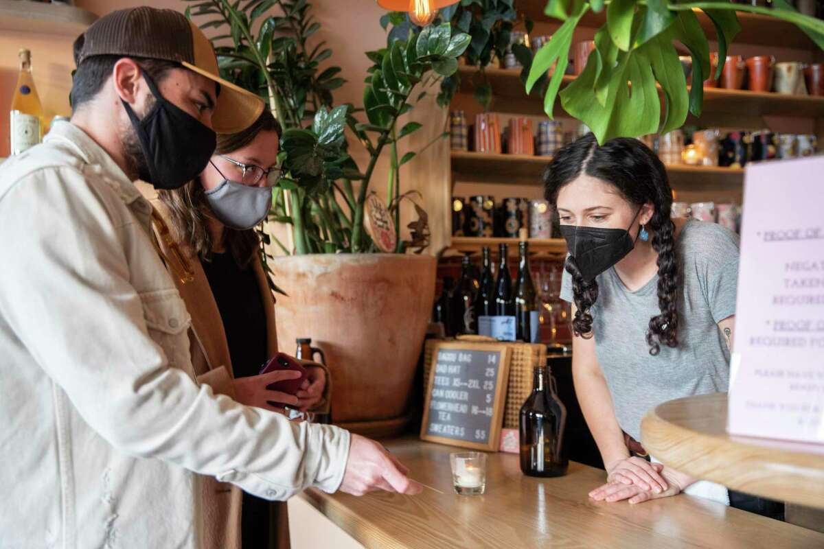Mark Kazour (left) and Isabel Kazour show their vaccine cards to Sierra Goldstein to sit indoors at Roses’ Taproom, in Oakland. California and most of the Bay Area are dropping their requirement that people wear face masks in indoor settings.