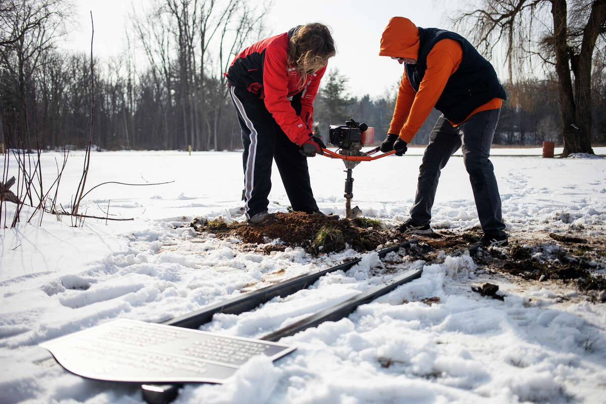 Drew Schmidt, left, and Daniel Davis, right, of MitchArt, dig two holes in the ground before placing a new sign in Porte Park Tuesday, Feb. 1, 2022, in honor of the families who were displaced by the 2020 dam failures.