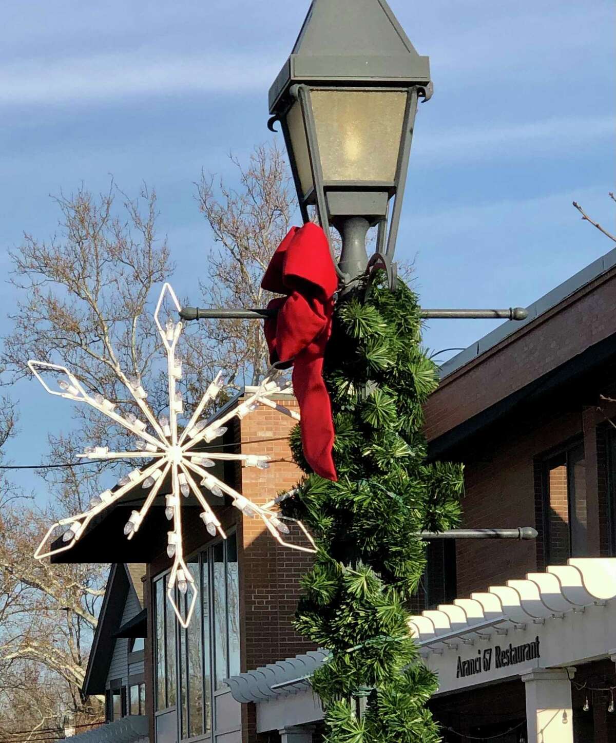 Members of the Making Wilton Beautiful initiative, and the Wilton Garden Club hope to make the town shine even brighter during the general holiday season, than it already is, and are thus launching a fundraising campaign to raise $8,400, to purchase an additional 24 snowflakes for the effort. A photo of a snowflake, a garland, and a bow, in Wilton, during a previous holiday season in the town, is shown.