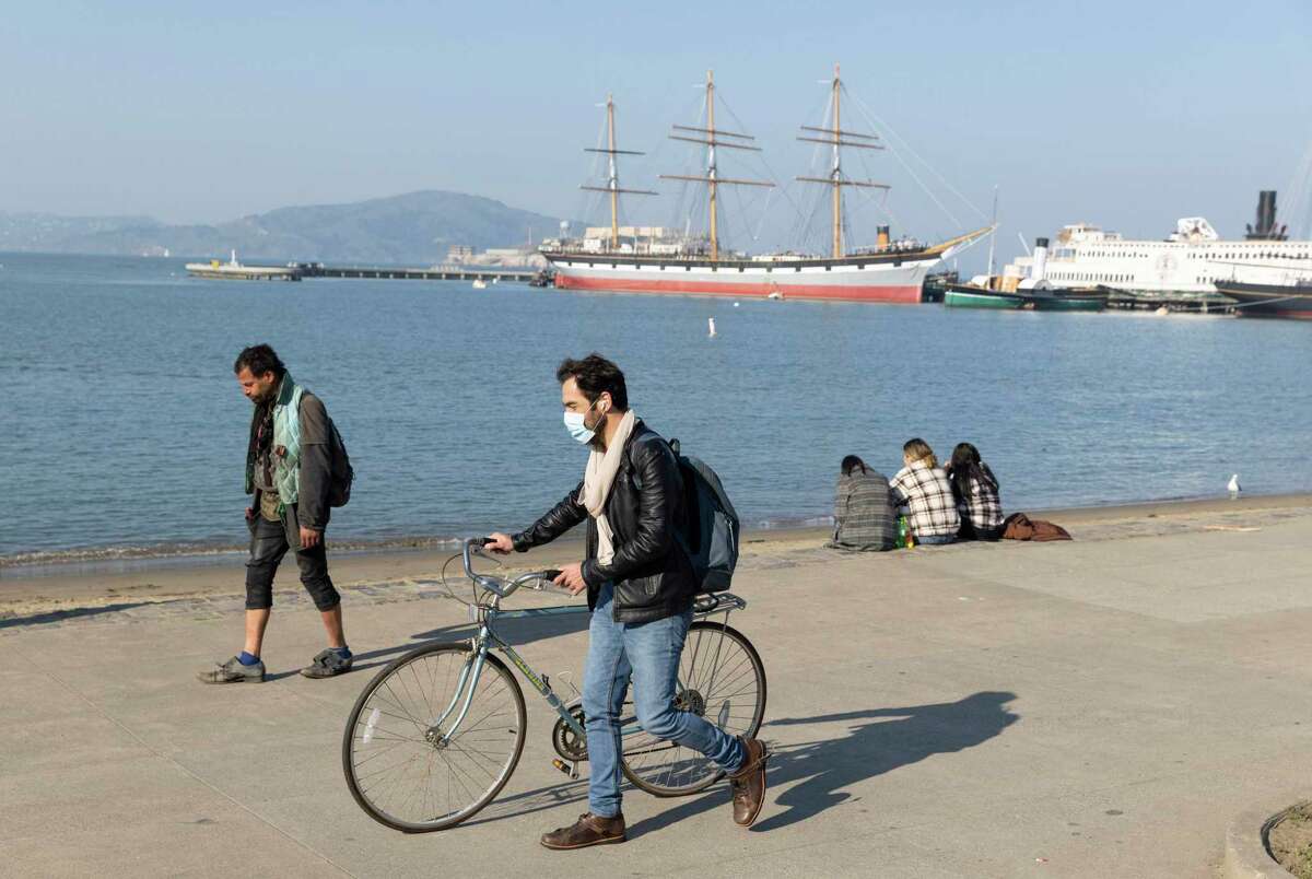 A cyclist in a mask walks through Aquatic Park in San Francisco. California is ending its universal mask mandate for indoor settings. Restrictions for the unvaccinated and in some settings will remain, however.