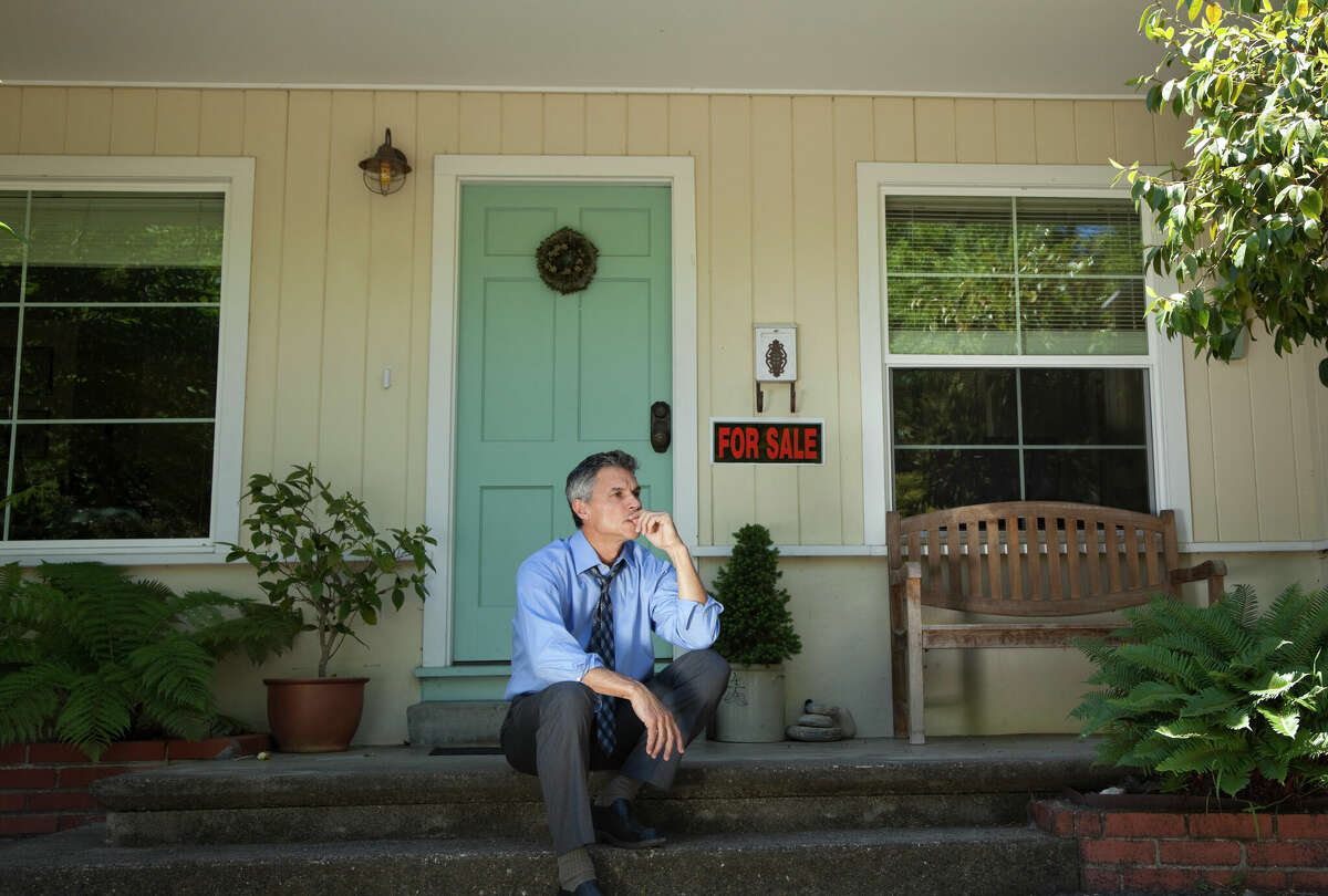 A man in a shirt and tie sitting on a doorstep in front of a home for sale. 