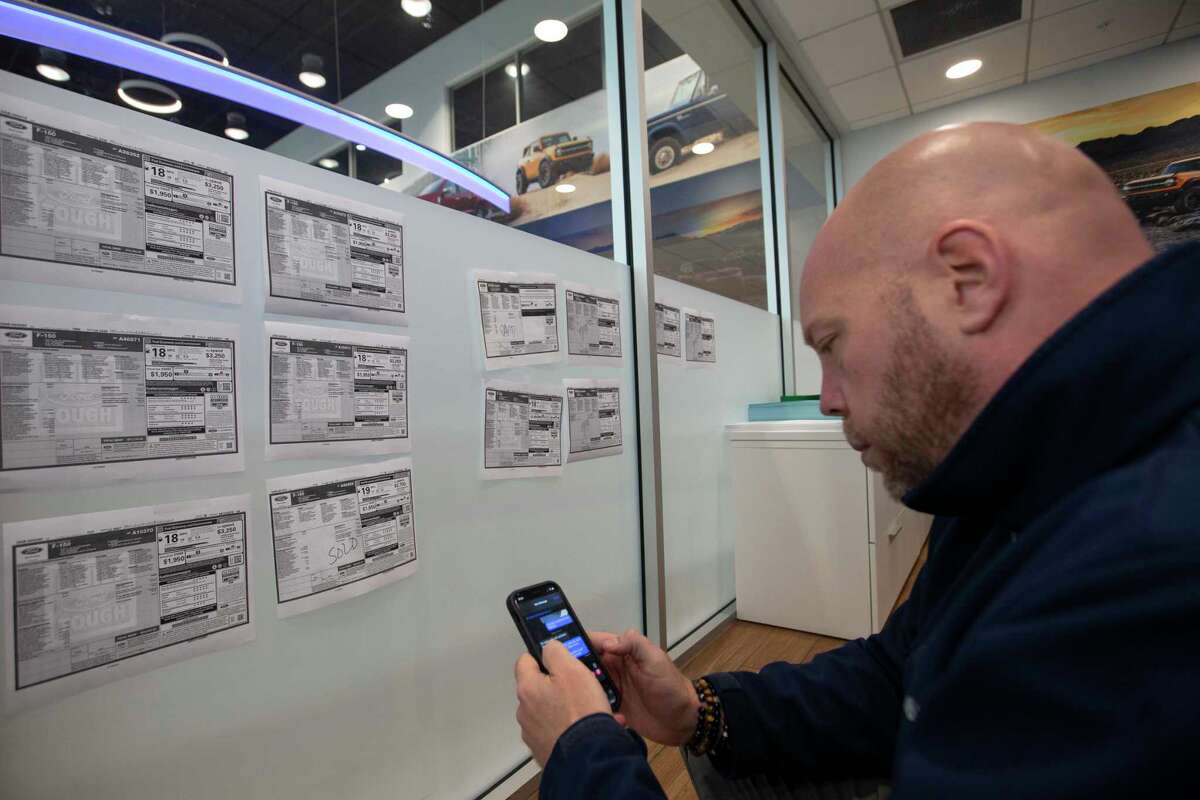Car sales specialist David Claar texts a customer, whom he has been working with for three months, about a F-150 truck that he might be interested and will be available at the dealership Thursday, Jan. 20, 2022, at Doggett Ford in Houston. The dealership puts window stickers of vehicles that will be delivered to the shop on the glass wall. Claar said half of the vehicles on the glass door would be sold before they are delivered.