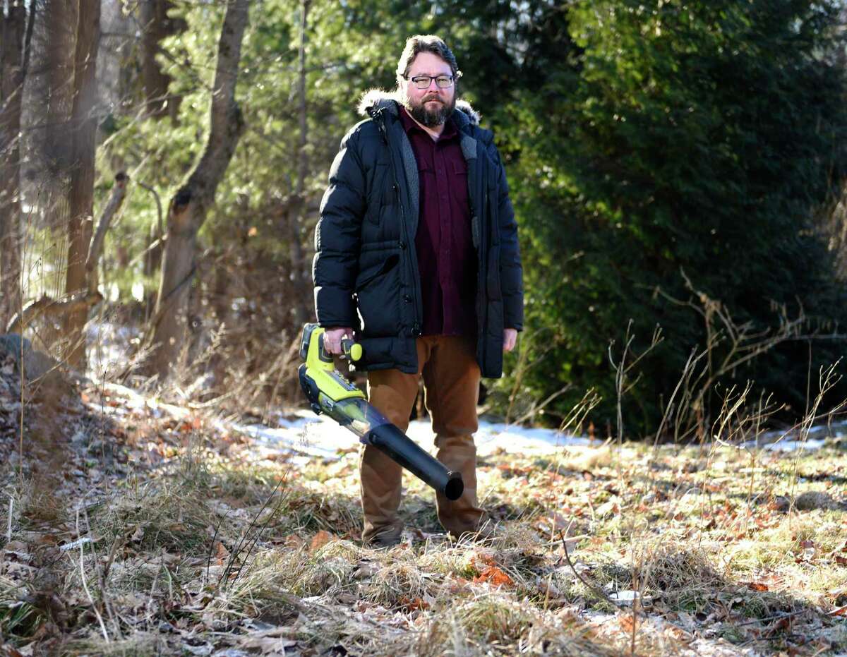 Jason Munshi-South poses with his electric leaf blower in a meadow of indigenous plants outside his home in Stamford, Conn. Thursday, Jan. 27, 2022. Munshi-South is bringing the anti-gas-powered leaf blower movement to Stamford, adding another city to the growing list of municipalities looking to restrict the machines. He has reduced his own use of lawn care services by defaulting to rakes and electric leaf blowers, but has also limited how much lawncare he actually needs by planting a meadow of indigenous plants in place of grass.