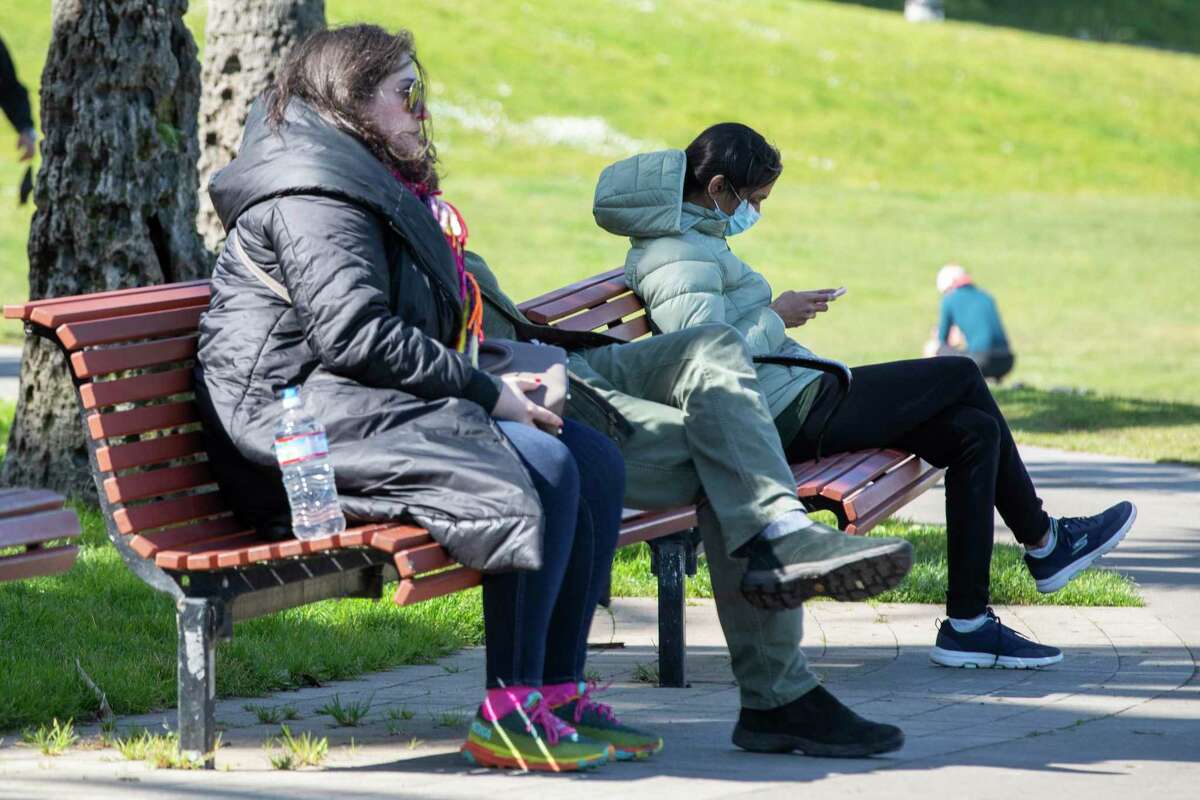 A person wears a mask while seated on a bench next to a mask-free person at Dolores Park in San Francisco, Calif. Friday, March 4, 2022.
