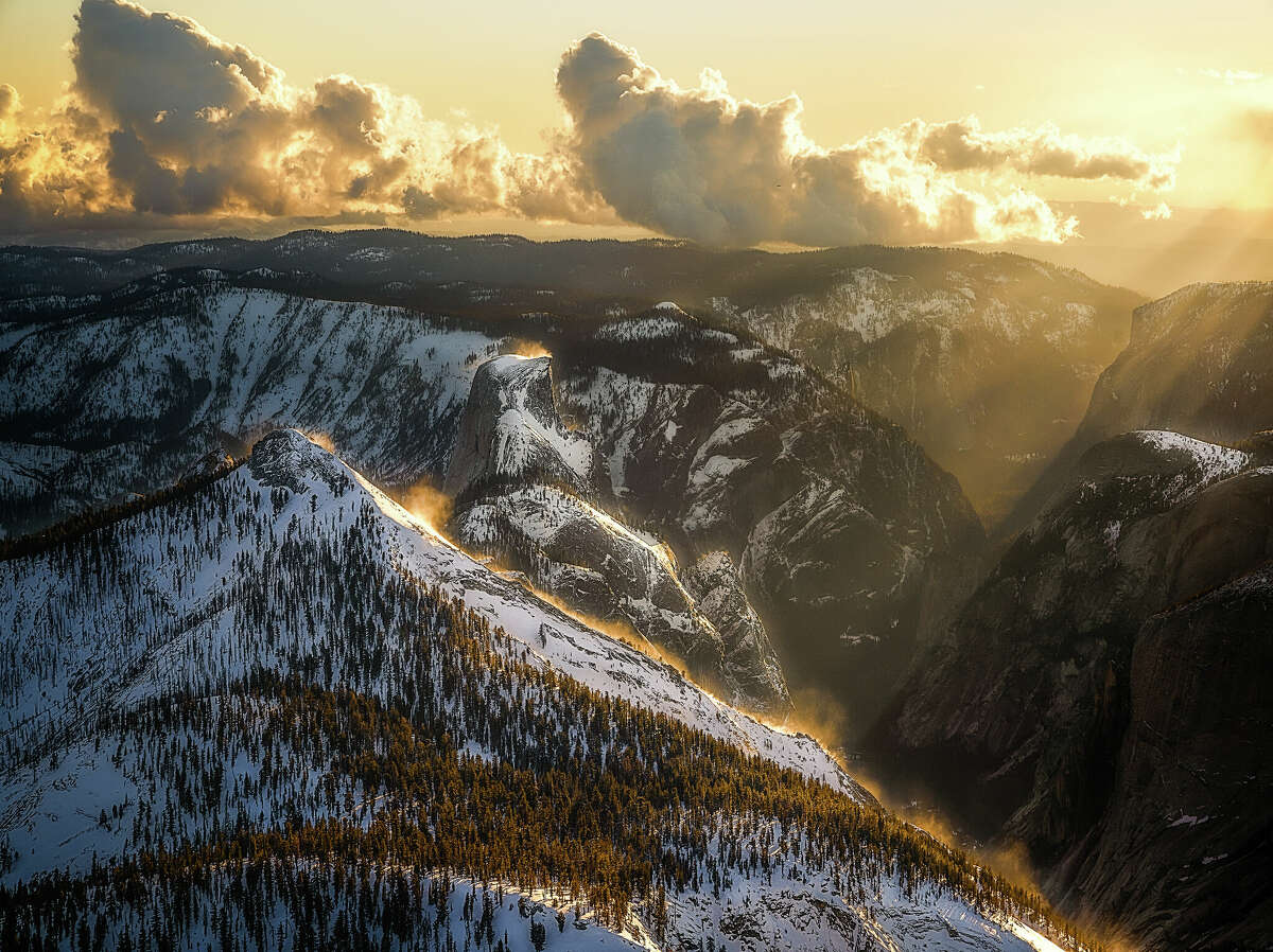 The sun's rays shine orange on blowing snow on top of a mountain in Yosemite National Park.