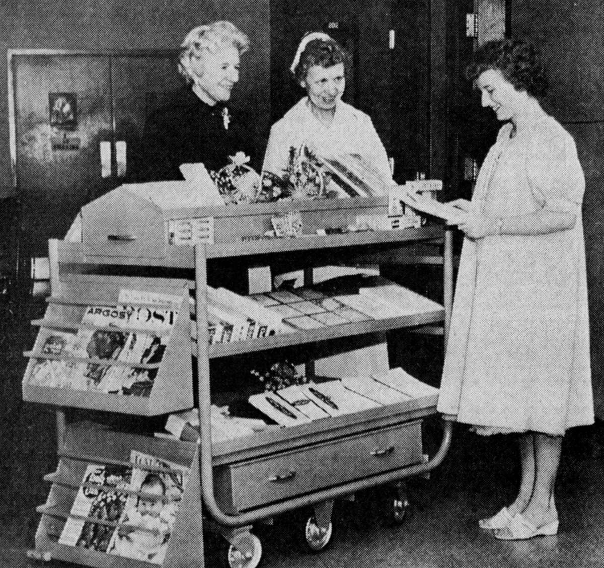 The Women's Auxiliary of Mercy-Community Hospital announces that the gift cart will soon be a familiar sight in the hospital corridors during visiting hours each day. Shown are Mrs. Alfred Kuechenmeister, Mrs. Ruth Shaw, registered nurse and Mrs. William Pearce, patient. The photo was published in the News Advocate on March 21, 1962.