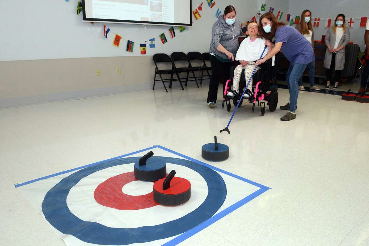 Physical therapists Kelly Hayes, right, and Tina Thayer help Emma Flannagan as she participates in curling during a Paralympics event at St. Vincent’s Special Needs Services, in Trumbull, Conn. March 18, 2022.