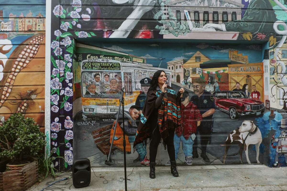 Lucía González Ippolito stands in front of her “Mission Makeover” mural on Balmy Alley, addressing Paseo Artistico event honoring her and other women in San Francisco’s Mission District.
