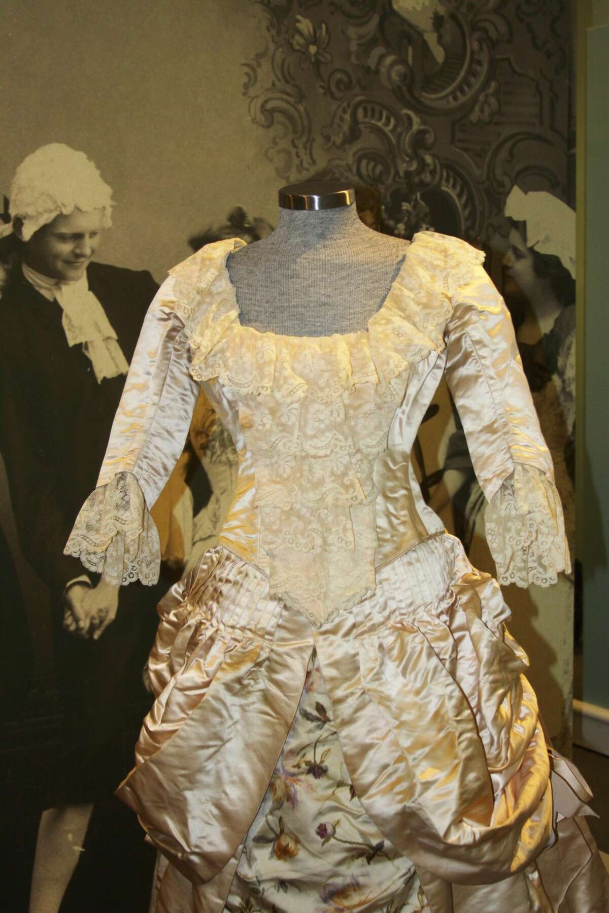 An elaborate gown of the Colonial Revival period in front of a tableau from that period.