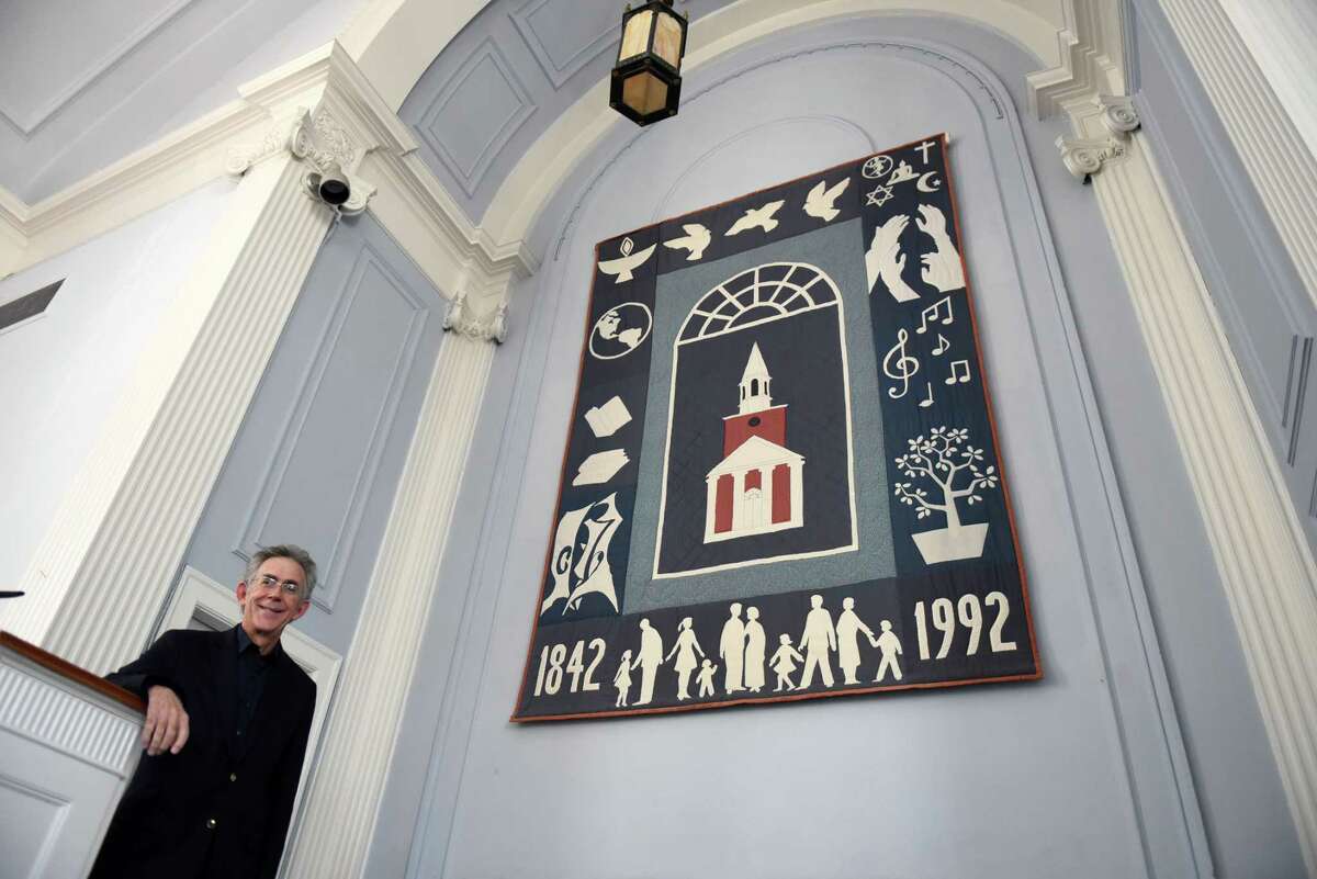 Rev Sam Trumbore of the First Unitarian Universalist Society of Albany stands in front the church’s alter quilt that was handmade by members of congregation and has been displayed in the front of the sanctuary for 30-years on Thursday, April 28, 2022, at the Albany Unitarian Universalist Church in Albany, N.Y. The quilt is to be replaced with imagery that better reflects the current congregation.