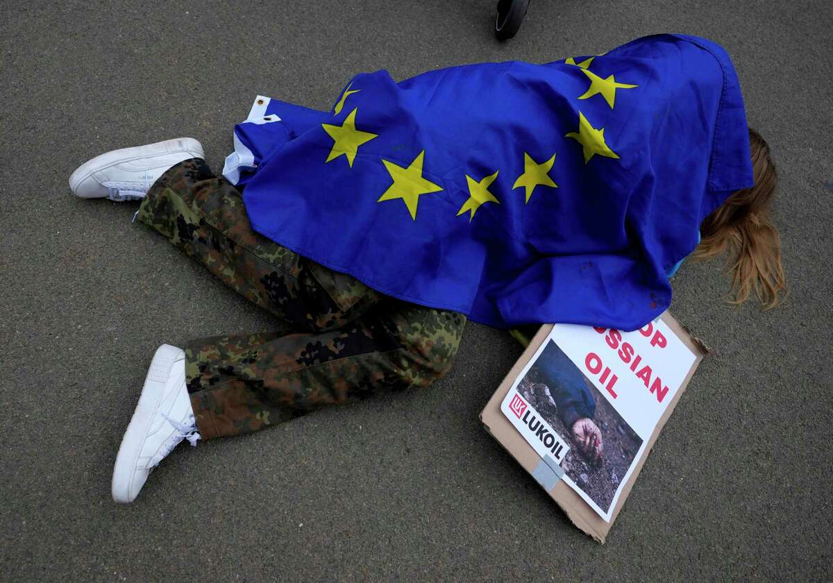 A protestor wearing military pants and covered by an EU flag takes part in a demonstration to call on the European Union to stop buying Russian oil and gas, outside EU headquarters in Brussels, Friday, April 29, 2022. Demonstrators, wearing Ukrainian flags and carrying boards laid on the ground, as they played a mock air siren, to represent the victims of the war. (AP Photo/Virginia Mayo)