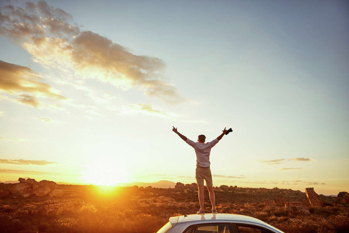 Rearview shot of a young man standing on top of a car with his arms outstretched in a rural landscape. 