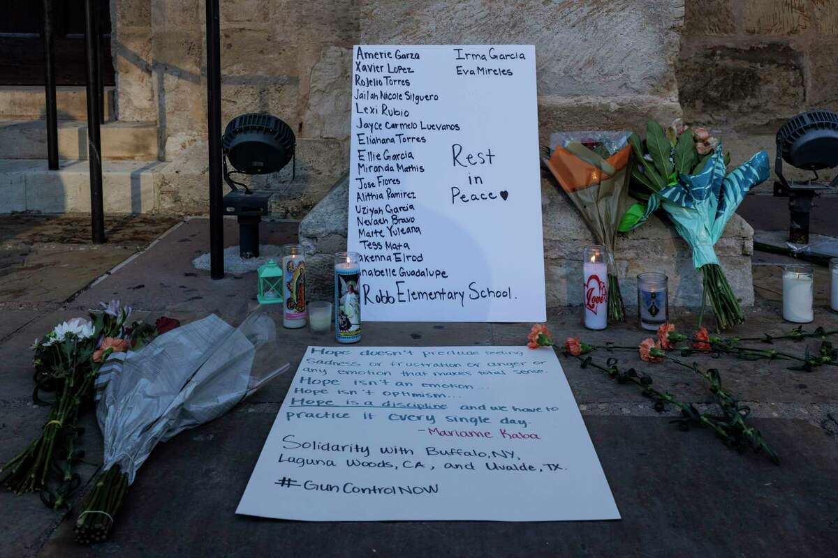 Flowers and candles are left Wednesday, May 25, 2022, outside San Fernando Cathedral in downtown San Antonio beside a sign listing the names of the 21 victims who died in the elementary school shooting Tuesday in Uvalde.