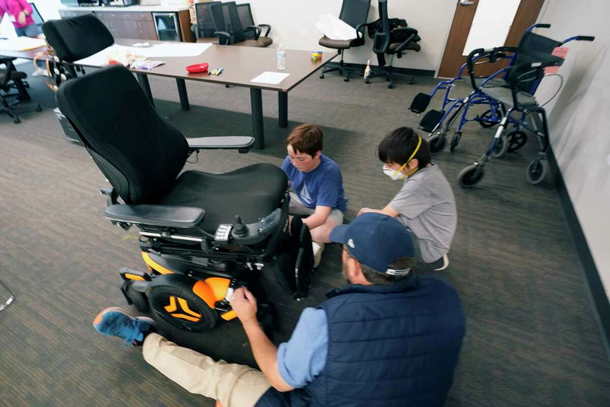 Tom Simon, front, goes over electric chair trouble shooting and repair with Charlie Warlick, left, and Alex Oliver during a workshop for young caregivers of ALS diagnosed family members in Dallas, Texas, Saturday, April 9, 2022. The children have gathered for a clinic to learn more about caring for people with Lou Gehrig's disease, or amyotrophic lateral sclerosis. It's a fatal illness that attacks nerve cells that control muscles throughout the body. (AP Photo/LM Otero)