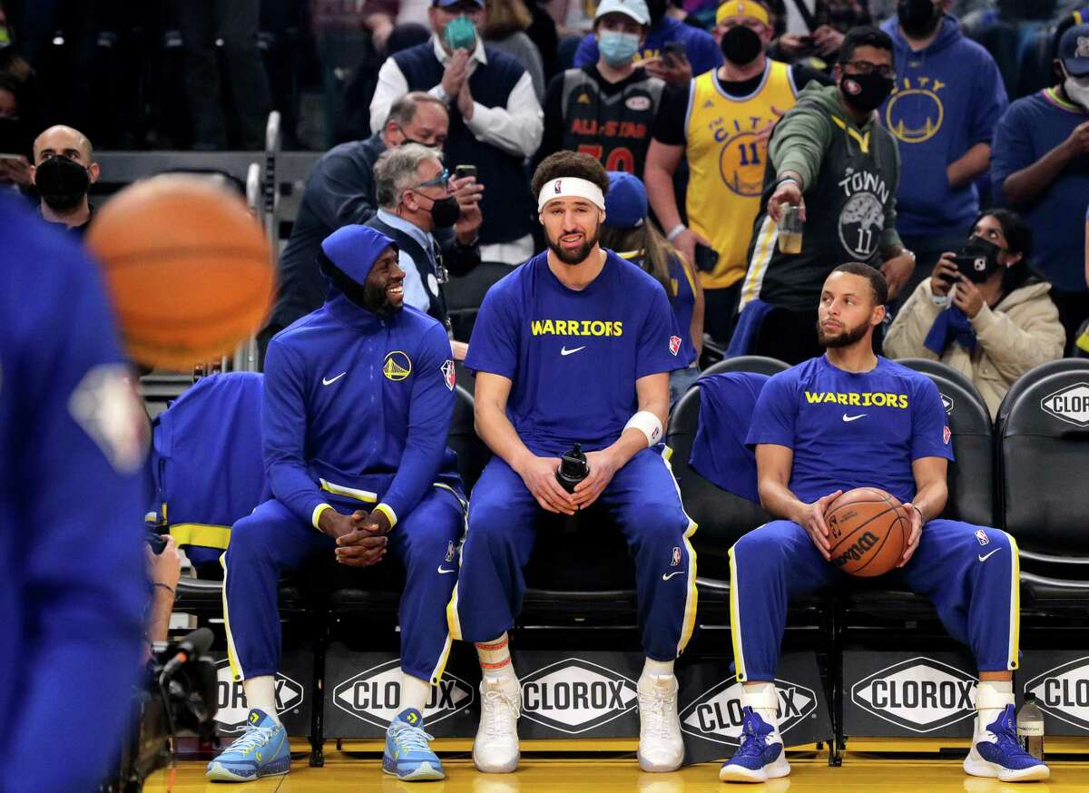 Klay Thompson (11) sits on the bench with long-time teammates Stephen Curry (30) and Draymond Green (23) before the Golden State Warriors played the Cleveland Cavaliers at Chase Center in San Francisco, Calif., on Sunday, January 9, 2022. Thompson returned to play after more than 900 days recovering from injuries.