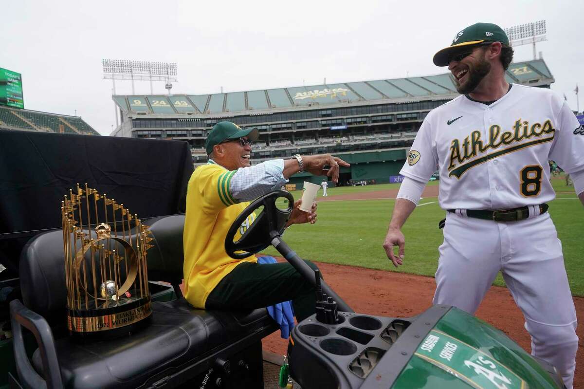 Reggie Jackson smiles toward Oakland Athletics second baseman Jed Lowrie (8) while sitting next to the World Series trophy during a celebration of the Athletics' 1972 championship team before a baseball game between the Athletics and the Boston Red Sox in Oakland, Calif., Saturday, June 4, 2022. (AP Photo/Jeff Chiu)