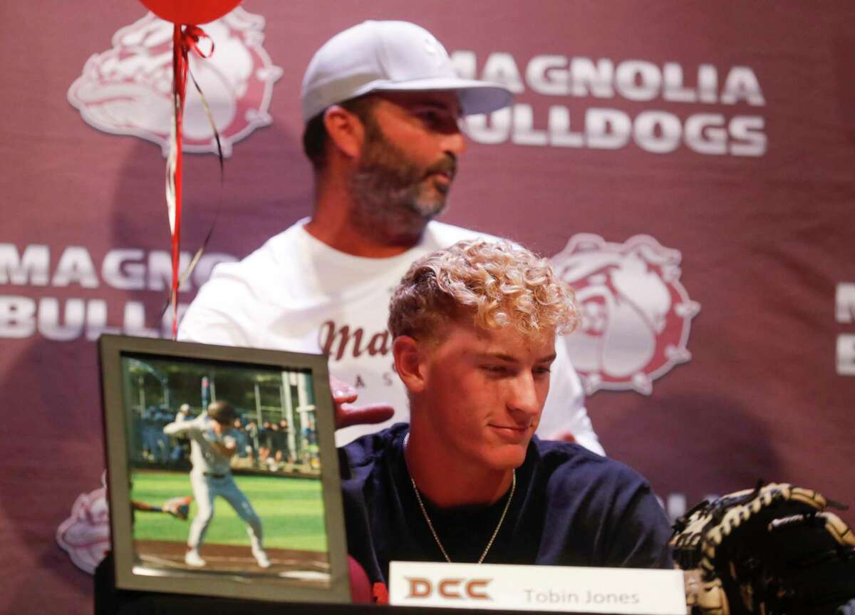 Magnolia coach Taylor Shiflett is seen in the background as Tobin Jones signed to play baseball for Dallas Christian University as he and eight other athletes from Magnolia High School signing to play sports at various colleges during a ceremony, Tuesday, May 10, 2022, in Magnolia.