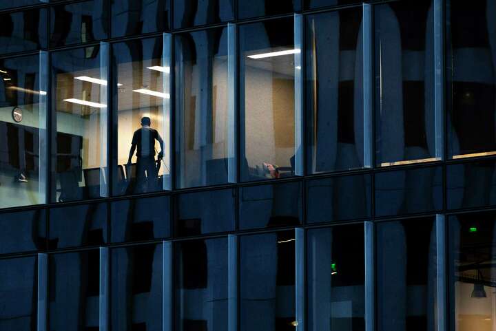 A worker is silhouetted in a lit room next to darkened windows of an office building seen from Salesforce Park.