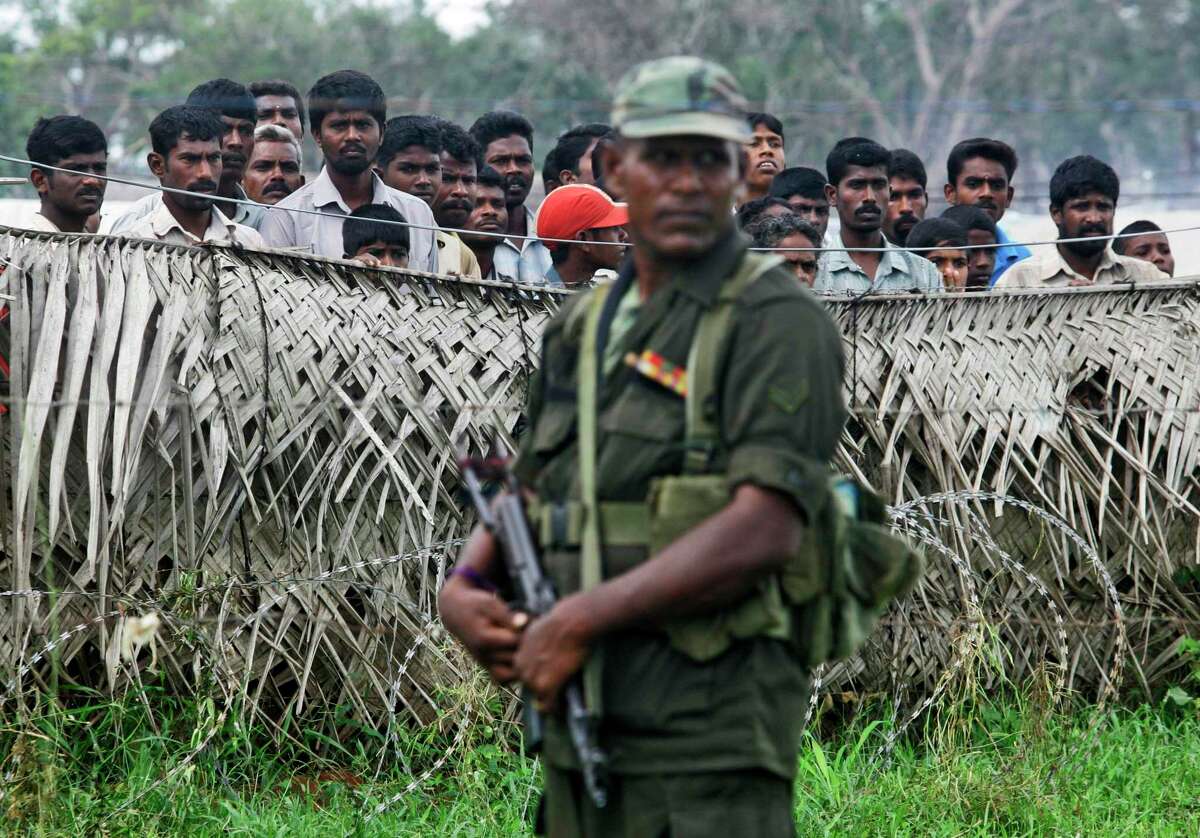 In this 2009 photo, a Sri Lankan army soldier stands guard next to a fence, as internally displaced civilians look on in the background at a camp for displaced in Vavuniya, Sri Lanka.