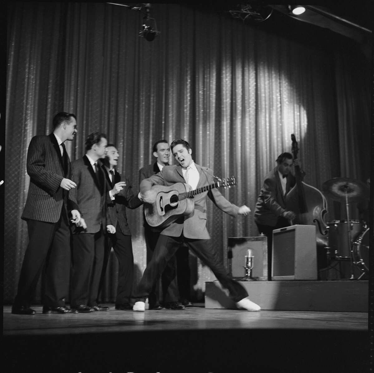 American rock and roll singer and actor Elvis Presley (1935 - 1977) dances as he sings during his second appearance on the Ed Sullivan Show, New York, New York, October 28, 1956. His back-ups singers, all in matching outfits, snap their fingers behind him, while and a bassist plays at the right. (Photo by CBS Photo Archive/Getty Images)