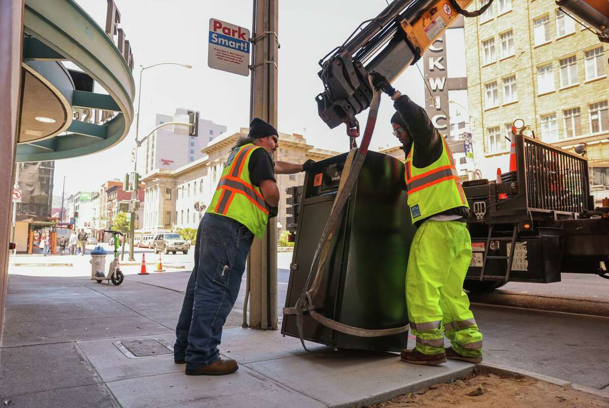 At Fifth and Mission streets, Mike Franov (left) and Ulysses Whittington with the Department of Public Works unload a bear saver model trash can, one of six models being tested.