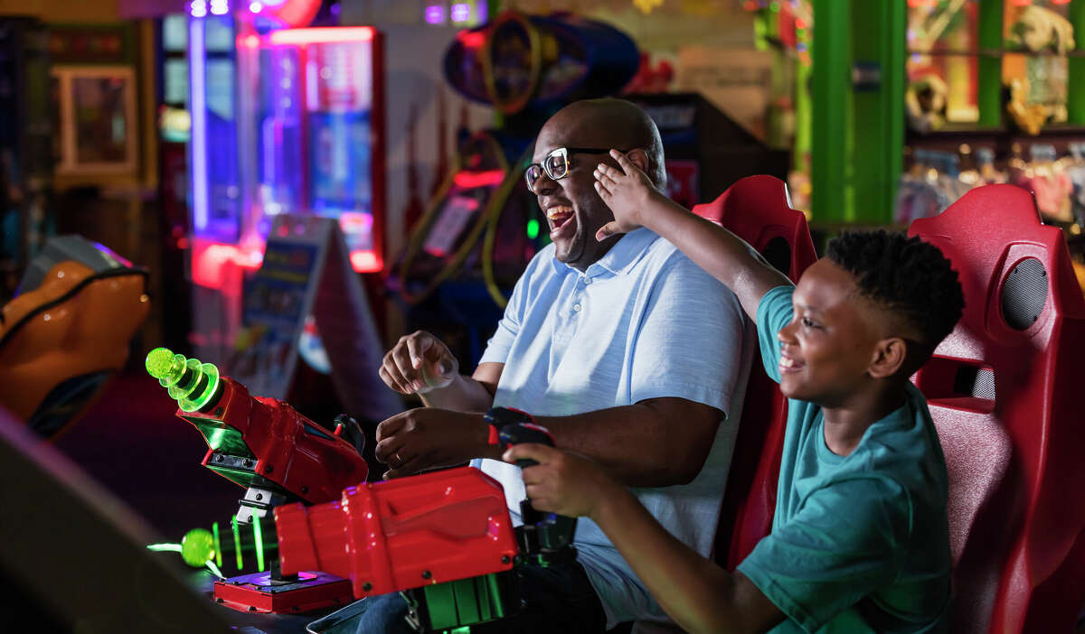 A father and son are having fun in an arcade, playing a game and pointing toy laser guns at a screen.