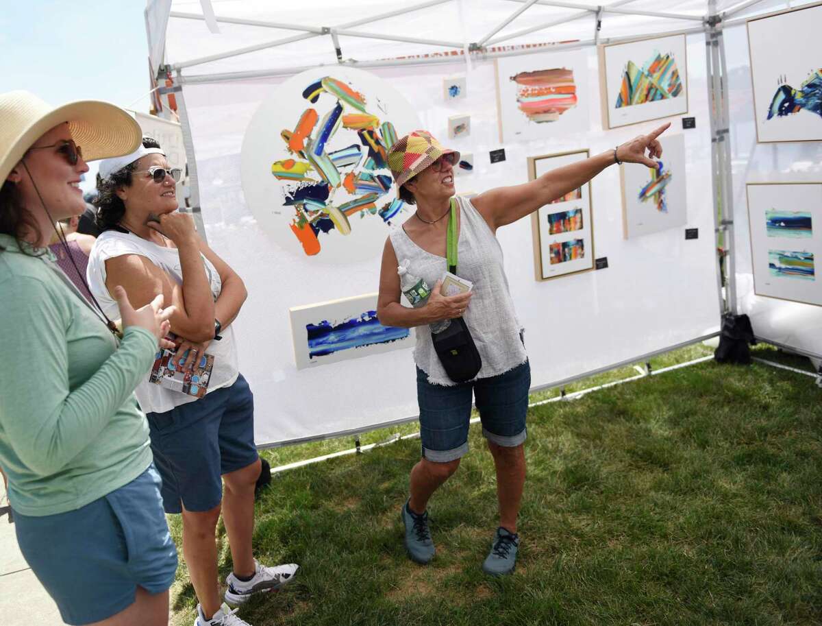 Artist Brooke Hirsheimer, left, of Little Dude Art, shows her work to Tricia Chapa, center, and Sandy Davis, of Austin, Texas, at the Stamford Arts Festival at Harbor Point in Stamford, Conn., Sunday, July 24, 2022. the seventh annual juried exhibition showcased one-of-a-kind works in a variety of mediums, including painting, sculpture, metalwork, printmaking, jewelry, and more.  The event included demonstrations, hands-on art projects and children's activities.