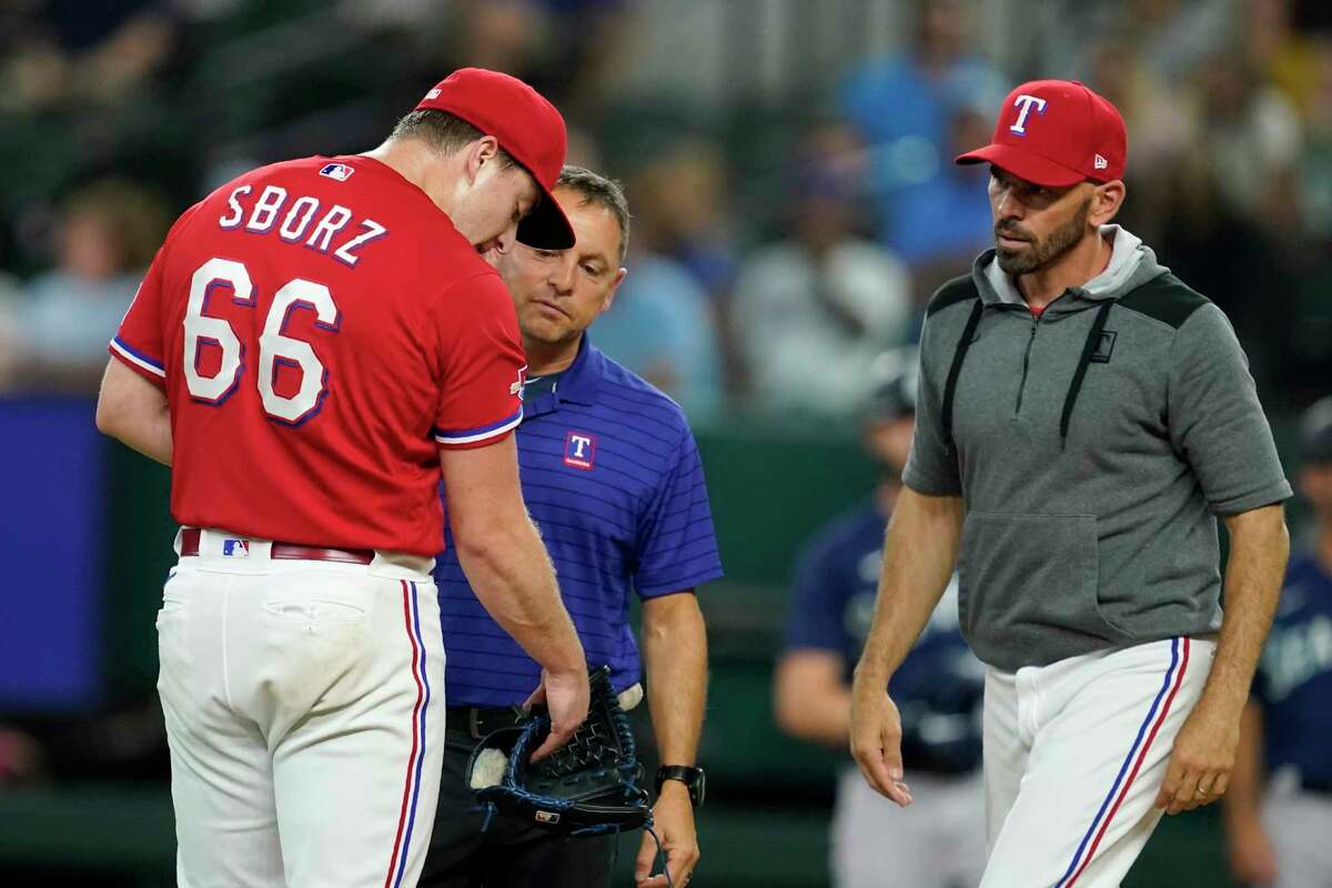 Texas Rangers starting pitcher Josh Sborz (66) is checked on by athletic trainer Matt Lucero, center, and manager Chris Woodward after Sborz was struck by the ball on a single by Seattle Mariners' Mitch Haniger during the first inning of a baseball game in Arlington, Texas, Friday, Aug. 12, 2022.