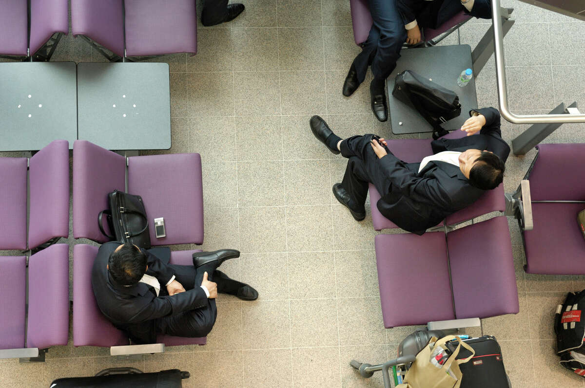 Business passengers with mobile phones and baggage wait sitting on purple seats at Manchester Airport.