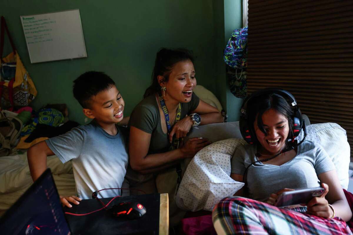 Daughter Angelika De Castro, 18, shows her mom Mary Jane De Castro (center), and brother Jericho De Castro, 9, that she can beat them at a video game on her phone in San Francisco, California on Monday, Aug. 22, 2022. Mary Jane is married with four children and a dog and they live in a studio apartment. Currently she is the only one working as her husband is suffering from a traumatic brain injury due to a motorcycle accident. Mary Jane’s days are long and she often doesn’t get a break.