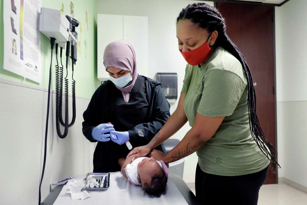 Nurse Wrood Hasan, left, administers an immunization shot to 2-month-old Kh’Alani Stewart, bottom, as mom Keaunni Sampract, right, supports the child at the Hope Clinic in Houston.