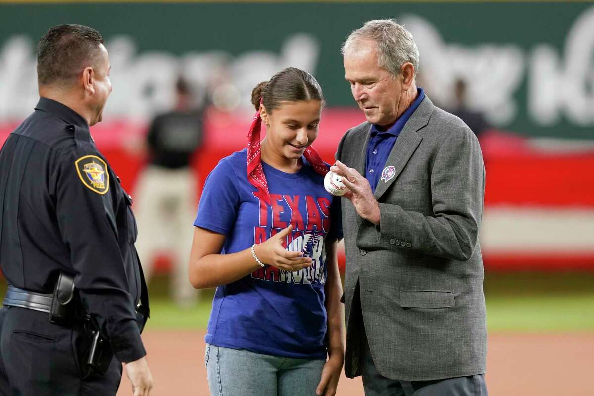 Former President George W. Bush, right, hands the ball to Andita Pollozani, center, for the ceremonial first pitch as her father, left, Fort Worth, Texas police officer Jimmy Pollozani, looks on to recognize the 21st anniversary of Patriot Day before a baseball game between the Toronto Blue Jays and the Texas Rangers in Arlington, Texas, Sunday, Sept. 11, 2022.