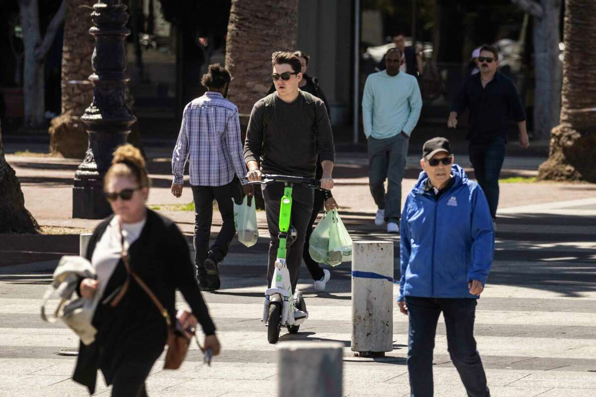 A scooter rider crosses an intersection on the Embarcadero in San Francisco. The city’s supervisors are asking for more citations to people who ride scooters on sidewalks.
