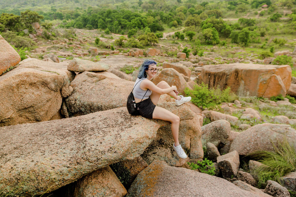 A young woman sits on a rock while hiking at Enchanted Rock State Natural Area in Texas. The park's Loop Trail is the only one to stay open after dark, inviting hikers to enjoy it with flashlights under a moonlit sky.