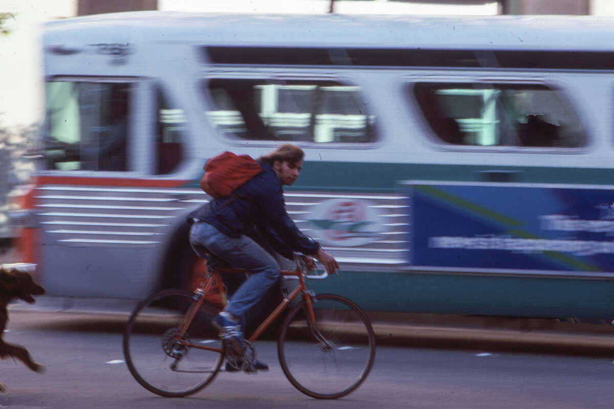 A man riding a bicycle past a bus with his dog right behind him. Date and place unknown.
