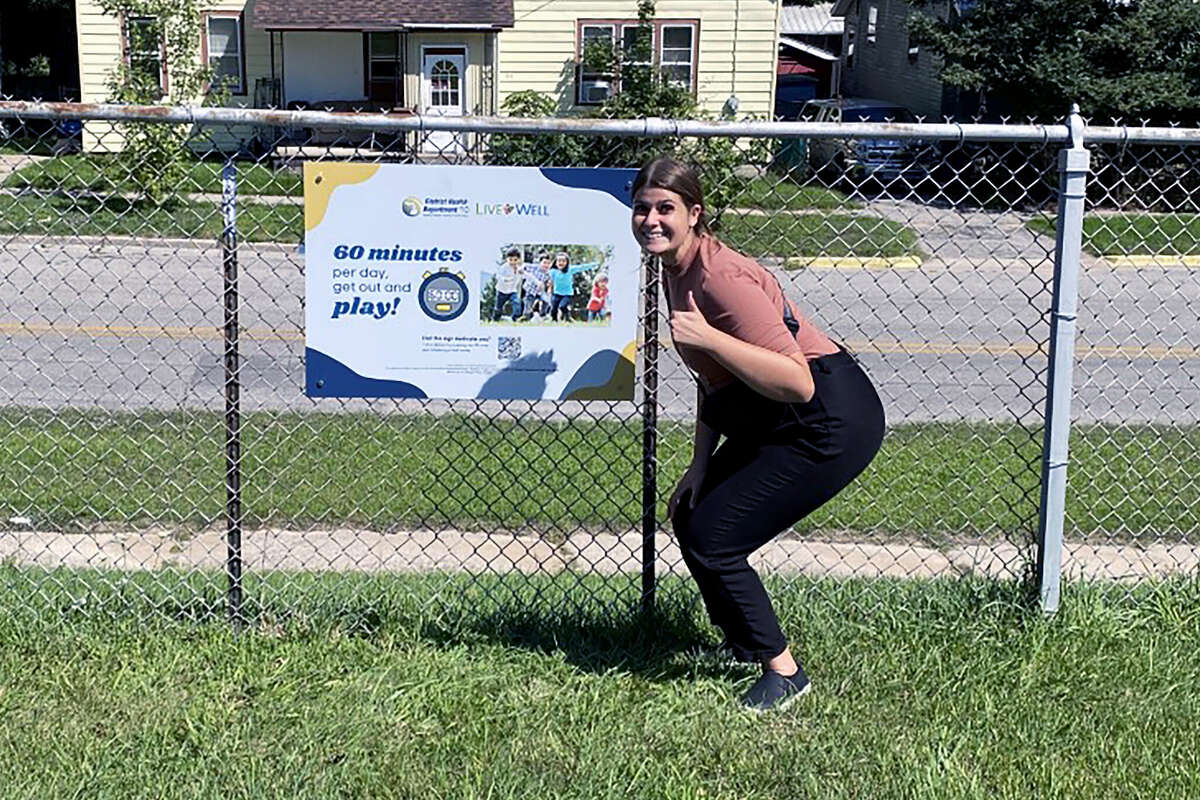 Kylie Davis poses near a sign in Maxwelltown promoting exercise and general wellness. Davis is the chair of the Live Well Manistee County coalition and is a health educator at District Health Department # 10.