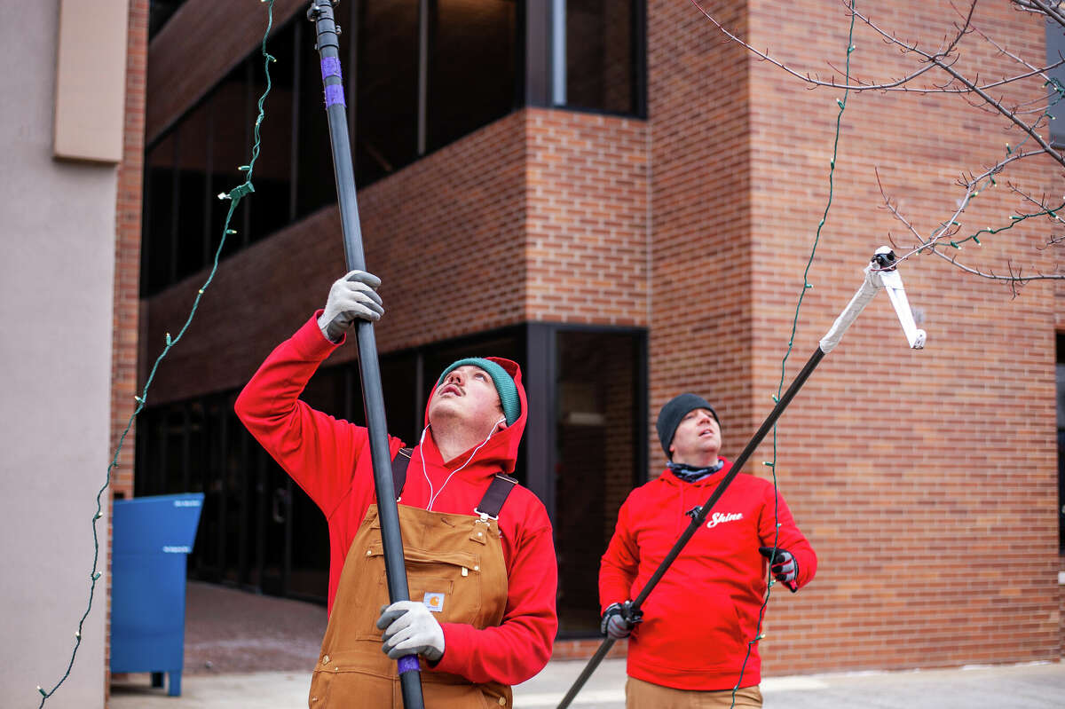 Nicki Hanson (left) and Jesse Dyer, technicians with Shine of Midland, wrap Christmas lights around trees on Nov. 29, 2022 in Downtown Midland. They are using a makeshift tool made from a window cleaner and a hook.