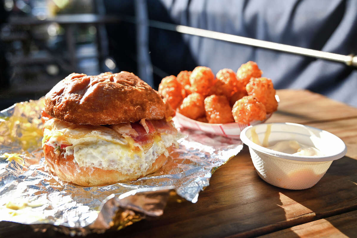 A bacon sandy made with 2 fried eggs, bacon, american cheese, pickled red onions, herby mayo, brioche bun with a side of tater tots and blowhole hot mayo aioli sauce.
