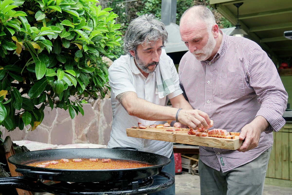 Jose Andres holds a tray with cooked belly fat (torrezno) as Diego Guerrero sets it on rice made in a paella pan in a scene from the Discovery + television series "Jose Andres and Family in Spain."