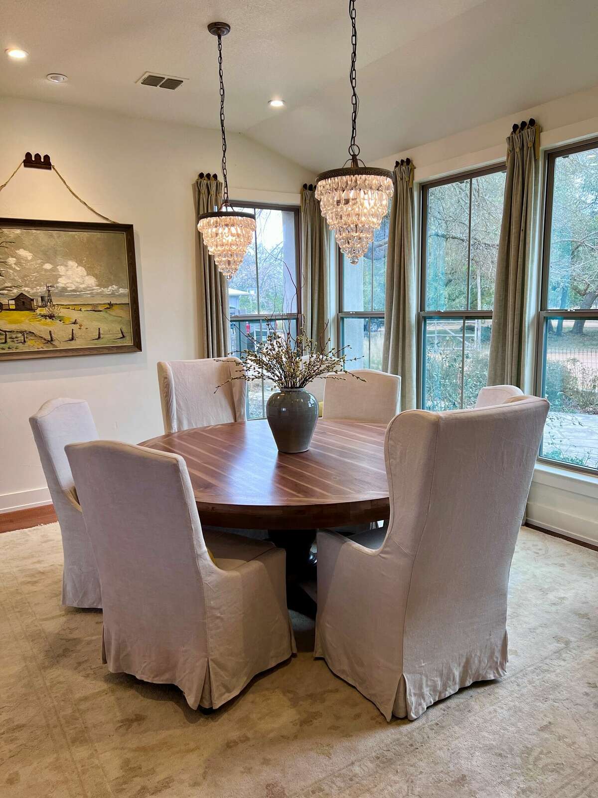 The elegant dining room has painted walls, curtained windows and two elegant chandeliers hanging above a round wood table surrounded by six chic skirted parson chairs.