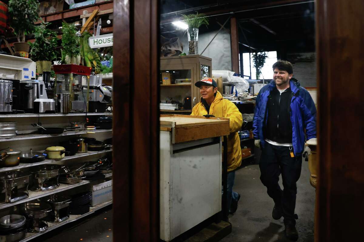 Jeff Dikio (left), Urban Ore worker, and Chris Amado, outside trade manager, are reflected in the mirror on a wardrobe as they transport a cart of cabinets to prepare items for delivery at Urban Ore.