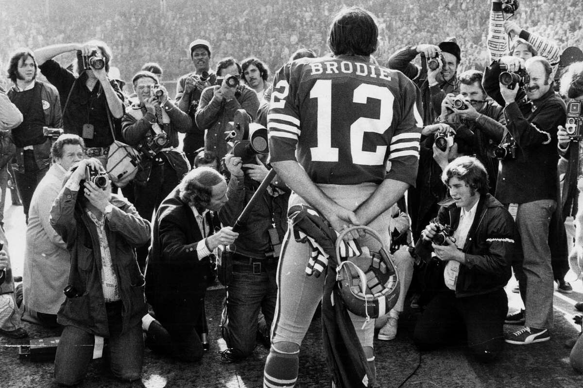 49ers quarterback John Brodie stands with his back to the camera wearing his number 12 jersey during a press conference, as several photographers in front of him take photos.