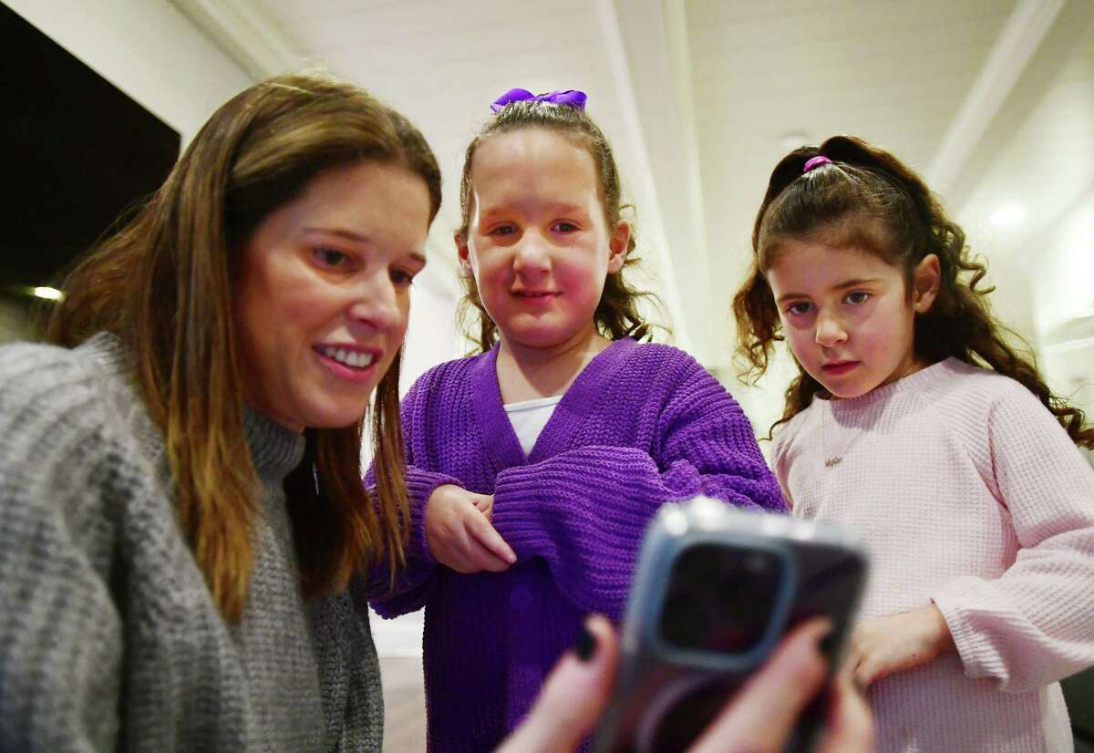 From left; Kimberly Greenberg watches a video on her phone with daughters Parker, 7, and Skyler, 4, at their home in Westport, Conn. on Wednesday, January 25, 2023. Parker suffers from a rare condition called HIST1H1E Syndrome. Greenberg is hosting the first annual Parker's Cure Bowling Bash charity event at Bowlero in Norwalk on Friday, January 27 at 8 pm.
