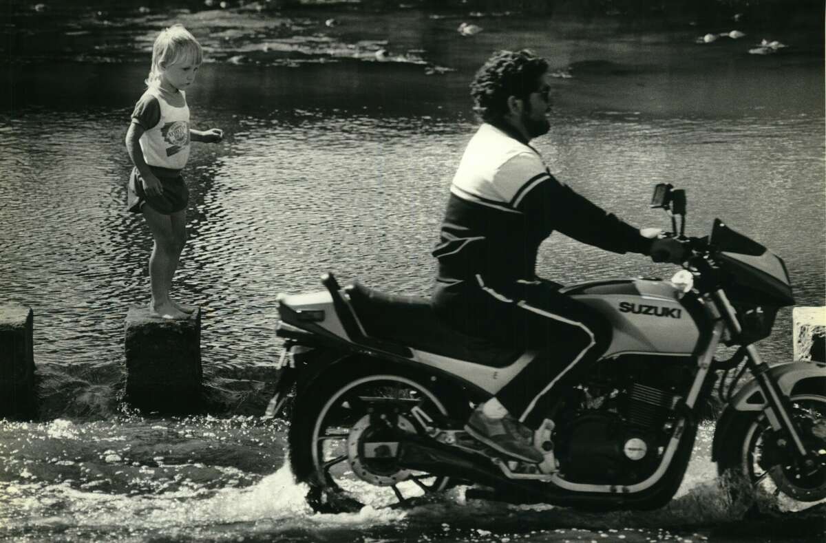 In this 1986 photo, 4-year-old Caleb Freese, walks on the stepping stones at the Brackenridge Park low-water crossing while a motorcyclist rides by.