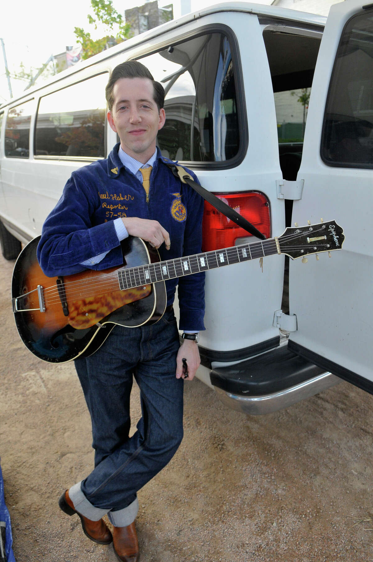 Pokey LaFarge wears an old fashioned haircut with cuffed jeans while he leans against a van holding his guitar.