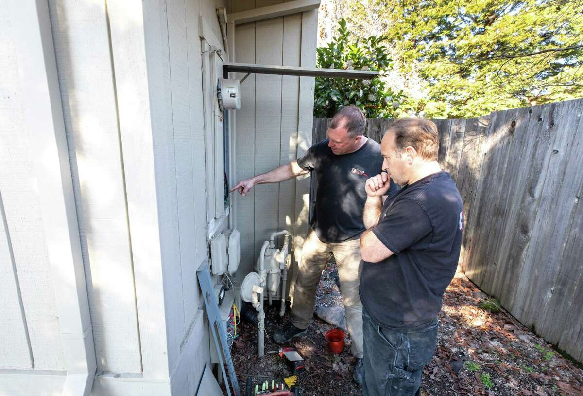 Thor Electrical employees Thor Cary (left) and Clint Petker work at the electrical panel and help install a heat pump at a home in Windsor.  Heat pumps can cost twice as much as gas stoves, several contractors said. 
