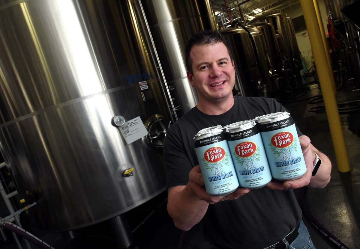Justin Gargano, CEO and founder of the Thimble Island Brewing Co. holds a six-pack of Thimble Island Brewing Co./Foxon Park White Birch Hard Soda in front of a 3,000-gallon vat containing the alcohol base for the hard soda, at the Thimble Island Brewing Co. in Branford.